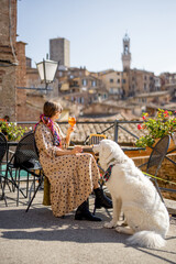 Naklejka premium Woman feeds her dog while sitting at restaurant with beautiful view on Siena town. Traveling Tuscany region in Italy. Concept of italian cuisine