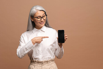 Amazed gray-haired woman standing isolated over beige background and pointing with finger on the smartphone for advertisement. Mockup image