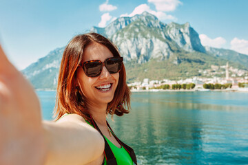 Face portrait of young cheerful woman taking selfie, wears sunglasses and green shirt in front of Lago Di Como lake at Italy. Holiday, travel, vacation, tourist, summer concept.