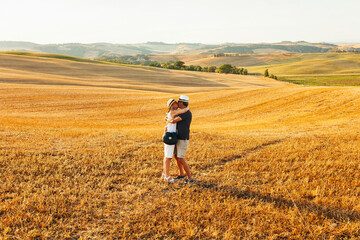Portrait of middle age couple kissing each other at yellow field during summer holidays at Tuscany, Italy. Senior couple wear stylish clothes and hat during sunset.