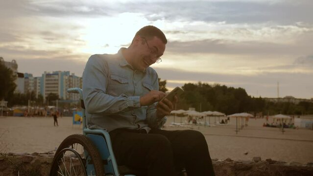 young man in a wheelchair is surprised and looking at his phone . Disabled guy in the background on the beach is holding the phone and laughing