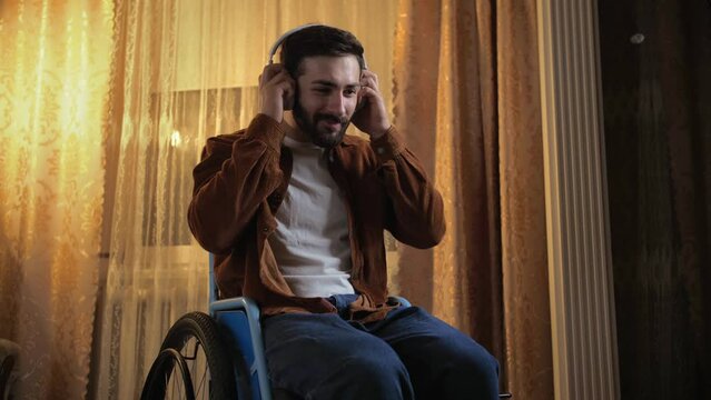 young man in a wheelchair puts on headphones and is surprised at how they sound. An disabled guy listens to music with headphones at home