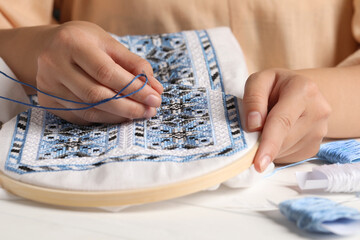 Woman embroidering shirt with blue thread at table, closeup. Ukrainian national clothes