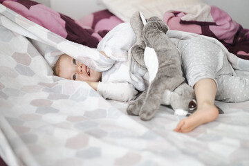 happy toddler girl covers herself with a blanket at home in bed with plush rabbit  - obrazy, fototapety, plakaty