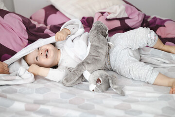 happy toddler girl covers herself with a blanket at home in bed with plush rabbit  - obrazy, fototapety, plakaty