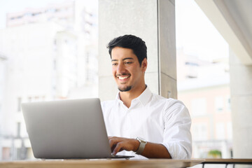 Latin male developer, freelancer, office employee is using laptop sitting at the desk oudoors. Young multiracial guy is typing on the keyboard, replying email, communicates with the customer