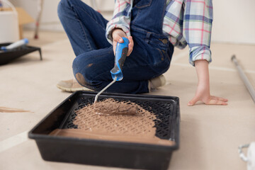 A child wearing denim overalls and a checkered shirt mixes brown paint with a paint roller. She is happy to help her parents with repairs at home.