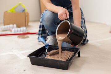 Obraz premium close-up of brown paint being poured into a plasma bowl. A bucket with paint is held by female hands. The girl is dressed in jeans, and in the background there are things that testify to the repair.