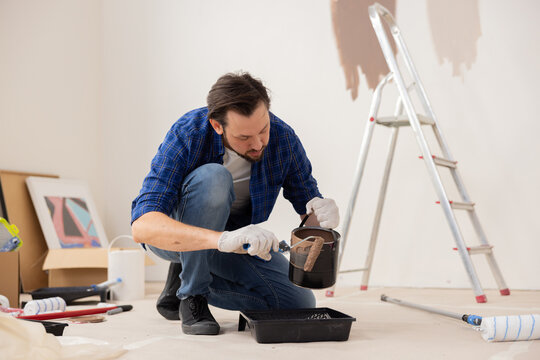 A 37-year-old man, wearing jeans, a white T-shirt and a checkered shirt, holds a can of brown paint and a paint roller in his hands. In the background, he has things that indicate renovation.