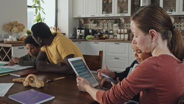 Low angle side view of young Caucasian woman sitting at table with little Black son on knees, boy using smartphone, mom using tablet computer