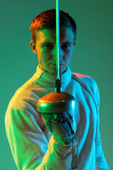Portrait of young tall male fencer in fencing costume and mask holding smallsword isolated on green...