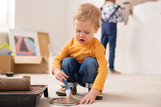 Close-up of a child, age 2, wearing jeans and an orange sweater, holding a paintbrush in his right hand. The child is very focused on drawing with a brush on the lid.