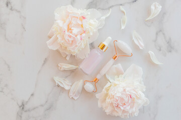 Jade face roller and essential oil for beauty facial massage therapy, Cosmetic tools for home treatment Face Serum or essence on white marble table with peony flowers and petals