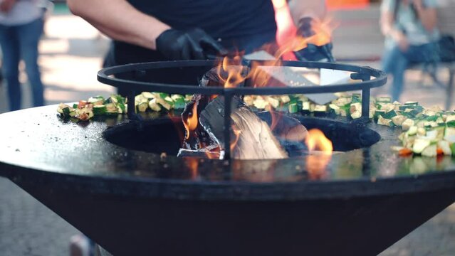 Chef roasting chopped zucchini, bell peppers and onion on traditional BBQ smoker stirring with cooking tongs, man grilling healthy vegan barbecue on hot charcoals, traditional barbecue festival and
