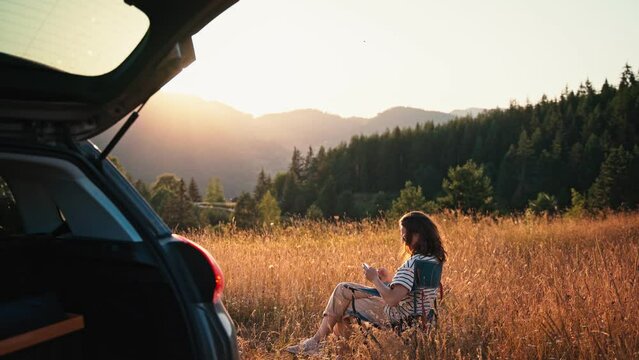 Cinematic shot of a young woman taking pictures with her smartphone while sitting in a folding chair during her solo car trip.