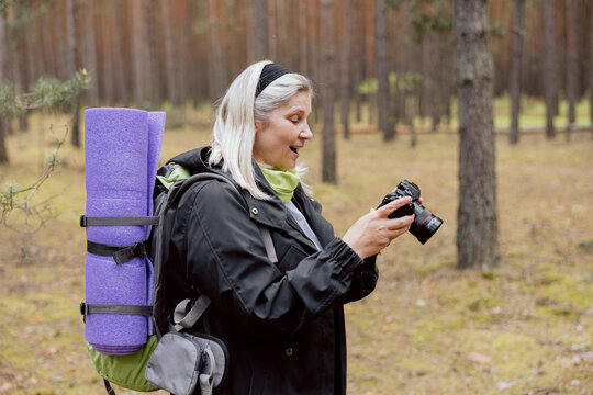 Surprised mother looking at camera screen seeing photos of her family. Elderly woman holding backpack and mat hikking camping trekking in the woods.