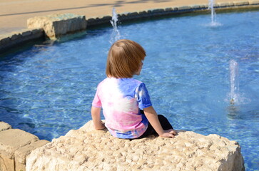 A small boy sits with his back to the camera. The child plays in the fountain. Summer holidays, sunny day