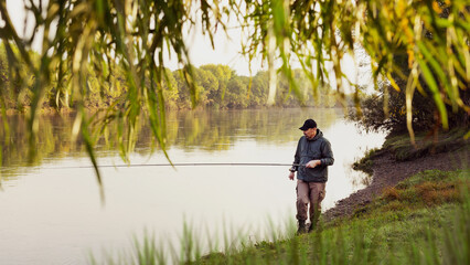 Fototapeta na wymiar A fisherman in a jacket with a fishing rod stands on the bank of the river. Fishing in the river. Fisherman with a fishing rod on the river bank. Fishing, spinning reel, fish.