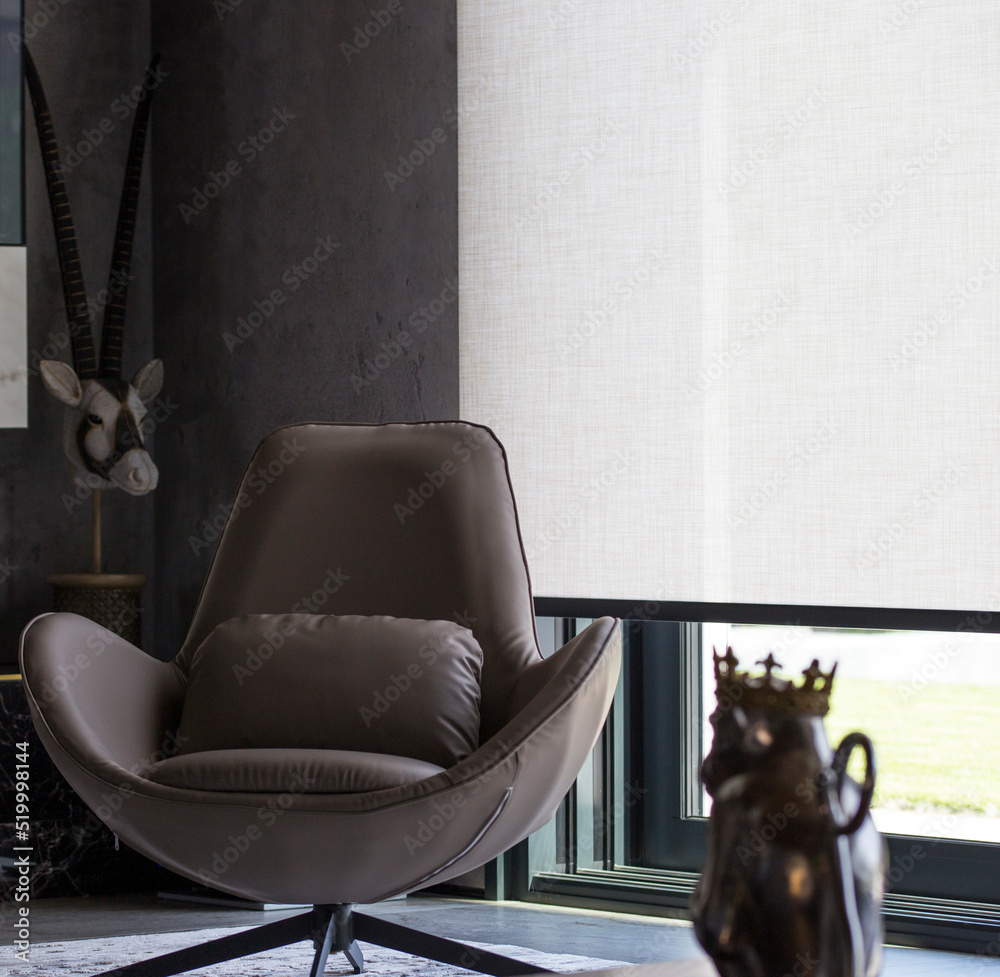 Aufkleber Roller blinds of large sizes on the window in the interior. Automatic solar shades, fabric with linen texture. In front of a large window is a chair on a carpet. Outside is a view of the garden.