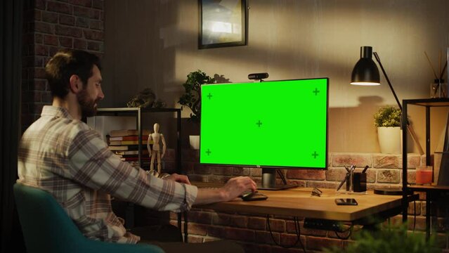 Young Attractive Man Working from Home on Desktop Computer with Green Screen Mock Up Display. Male Checking Corporate Accounts, Messaging Colleagues. Loft Living Room in the Evening. Zoom In Shot.