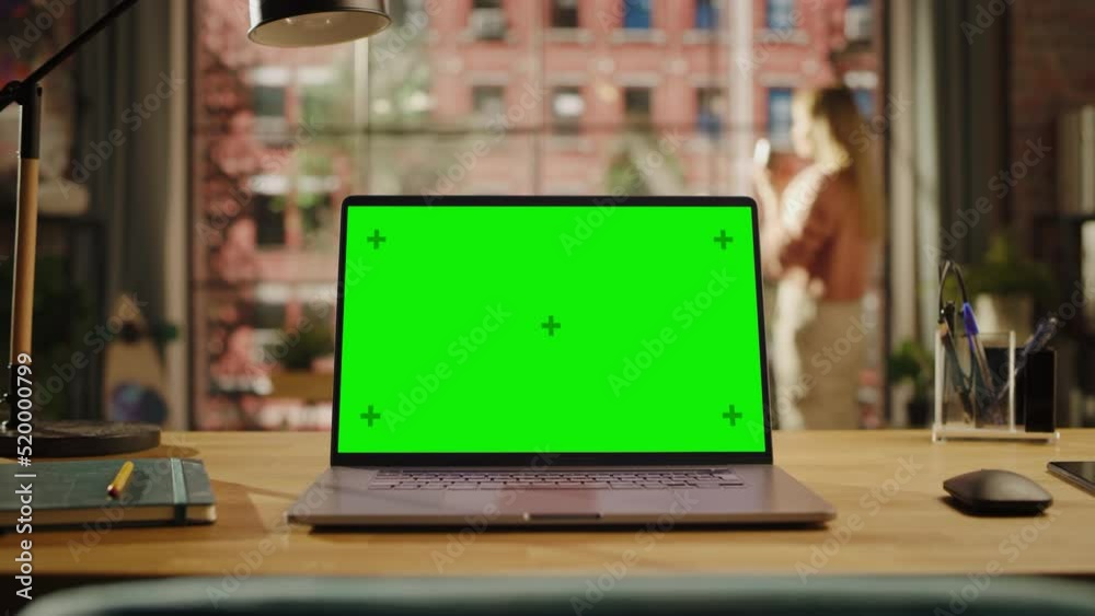 Poster Laptop Computer Display with Mock Up Green Screen Standing on a Wooden Desk in Living Room. Chroma Key Monitor at Home or Creative Loft Office with Female Looking Out of Window in Background.