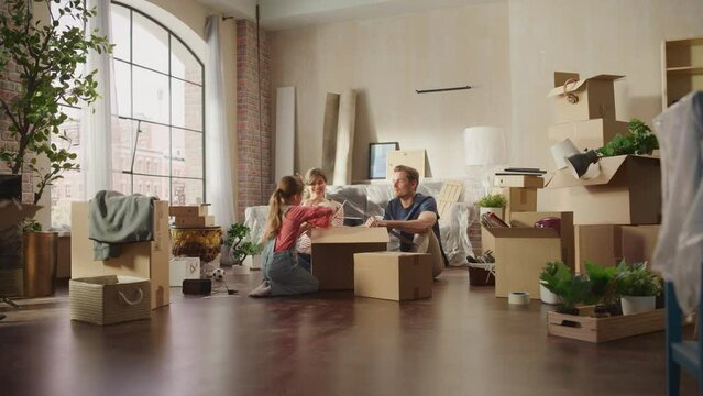 Happy Homeowners Moving In: Lovely Couple Sitting on the Floor of Cozy Apartment Unpacking Cardboard Boxes, Little Daughter Joins them. Cheerful Day, Harmony, Happiness, Sweet Home for Young Family