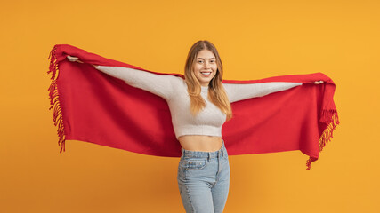 A young girl, spreading her arms wide, smiling broadly, demonstrates a red scarf, on a yellow background.