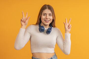 A young girl with long hair, with blue headphones around her neck, shows a peace sign, with both...