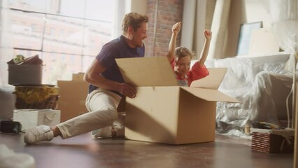 Moving in: Young Family of New Homeowners Has Fun. Father Driving Lovely Little Daughter in Cardboard Box. Dad and Pretty Cheerful Child Racing Together Through the Sunny Apartment Living Room. 