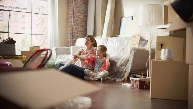 Moving in: Happy Mother and Daughter Talk, Sitting on a Living Room floor of New Cozy Home. Cheerful Young Family, Dream, Imagine Good Times in Apartment. Unpacked Cardboard Boxes around. Slow Motion