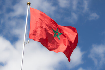 Fototapeta na wymiar The flag of Morocco flies against a clear blue sky with white clouds. Close-up, perfect for news.