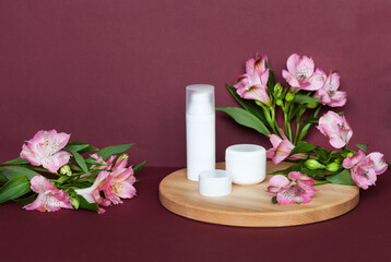 Presentation of summer cosmetic products: anti-aging creams for the face, eyelids and moisturizing gel with vitamin extract on a wooden podium and a burgundy background with alstroemeria flowers