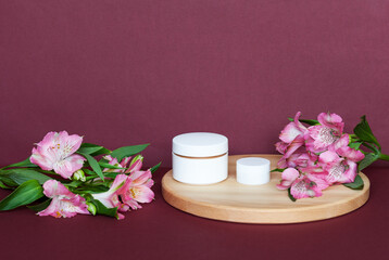 Natural rejuvenating summer cream for face and eyelids with floral extract on wooden podium and fresh pink alstroemeria flowers on burgundy background. Cosmetic products presentation concept