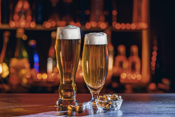 Glass of beer on bar counter. Jugs, mugs, pints of brew beverage, ale, cider on wooden table in pub, bar. Backlit dark room, showcase with craft beer bottles in brewery. Salted pistachio nuts snack