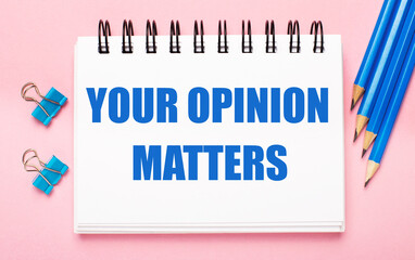 On a light pink background, light blue pencils, paper clips and a white notebook with the text YOUR OPINION MATTERS