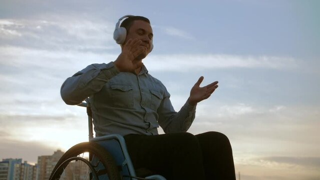 young man in a wheelchair wearing headphones listens to music and dances to it.Disabled guy in the backdrop of a beach in good mood dances while sitting in a wheelchair.