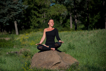 young beautiful woman doing yoga exercises on the stone among green grass in the park in sunny day 