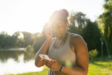 Plus sized African American woman using mobile phone and earphones before exercising at the park in a  summer day