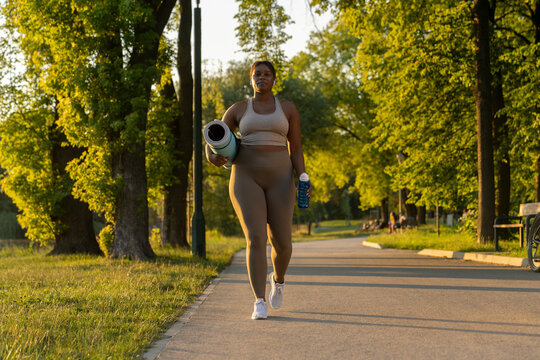 Plus sized African American woman walking with exercise mat and water through the park in a summer day