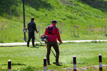 Two men cuts the grass in the garden with a hand-held electric lawnmower.	