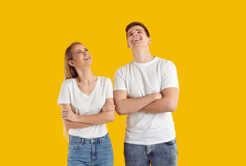 Happy young couple dreaming or thinking about something. Joyful man and woman in white tee shirts and blue jeans standing with their arms crossed isolated on yellow background, looking up and smiling