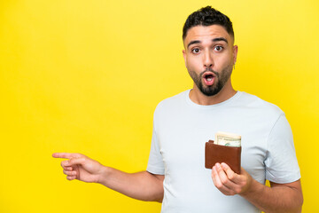 Young Arab man holding a wallet isolated on yellow background surprised and pointing side