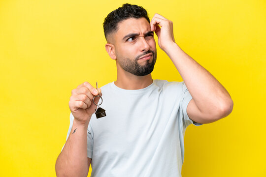 Young Arab man holding home keys isolated on yellow background having doubts and with confuse face expression