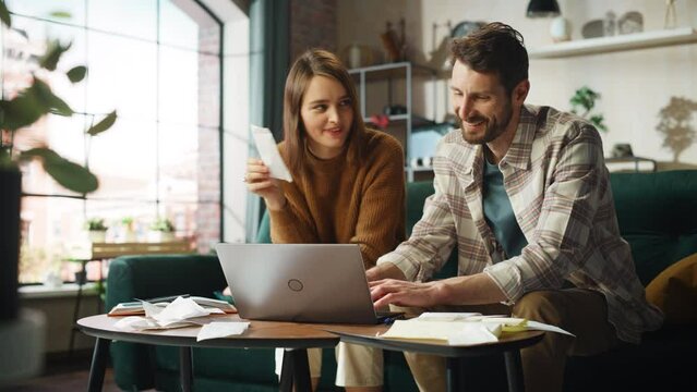 Doing Accounting at Home: Happy Couple Using Laptop Computer, Sitting on Sofa in Apartment. Young Family Filling Tax Forms, Mortgage Documents, Bills, Checks, Balances, Invoices are in Order