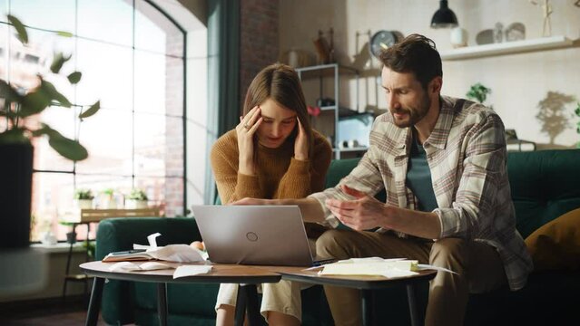 Stressful Dramatic Accounting at Home: Couple Using Laptop Computer, Sitting on Sofa in Apartment. Young Family Filling Tax Forms, Mortgage Documents, Bills, Checks, Balances, Invoices are in Order