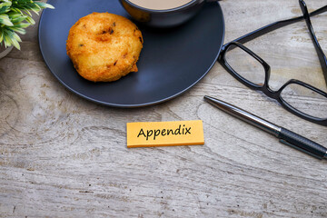 Word Appendix on the orange color post-it note on the wooden office table with a pen, a cup of coffee, cookies, and eyeglasses