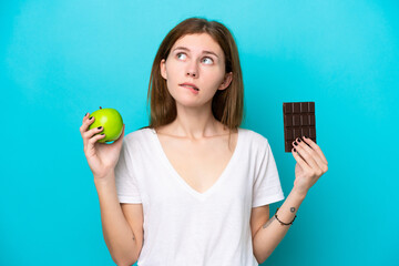 Young English woman isolated on blue background having doubts while taking a chocolate tablet in...