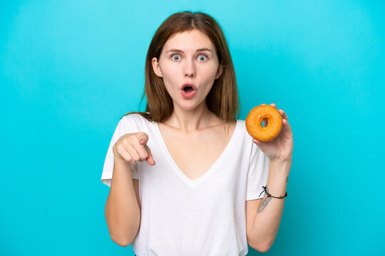 Young English woman holding a donut over isolated blue background surprised and pointing front