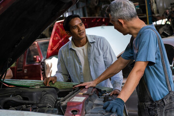 Fototapeta na wymiar senior Asian mechanic looks for parts to fix a broken engine and stands in front of the car feeling tired talking wiht man about engine broken in the garage.