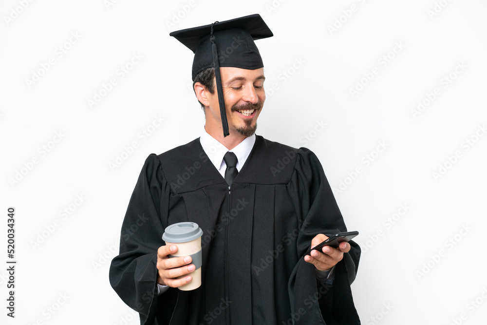 Wall mural Young university graduate man isolated on white background holding coffee to take away and a mobile